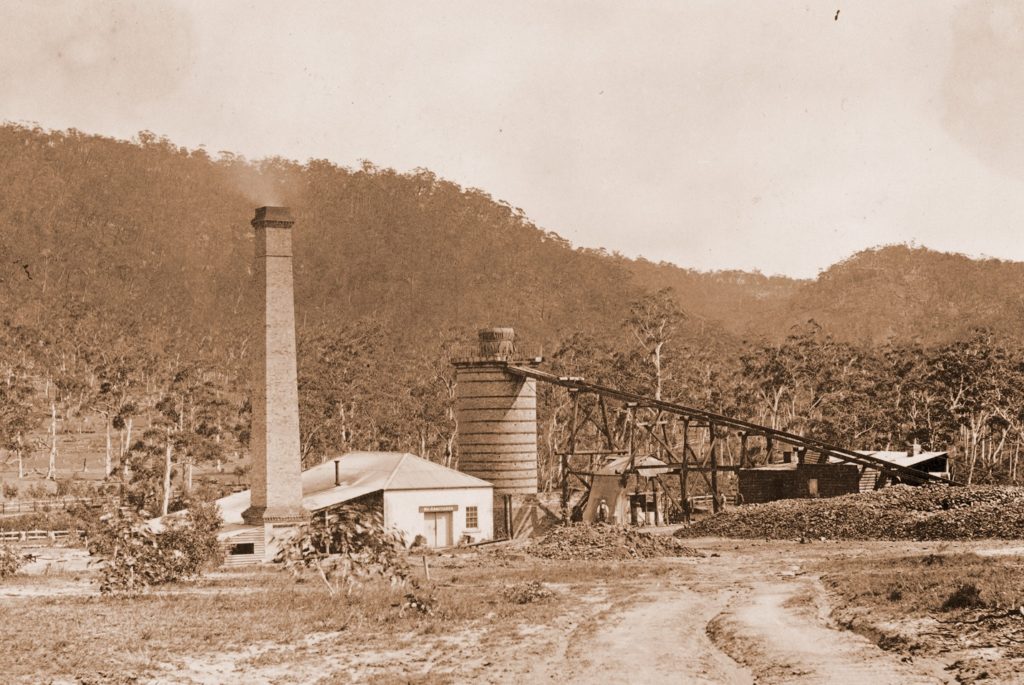 Sepia photograph showing a tall brick chimney attached to a small house and surrounded by Australian bushland. To the right is a large cylindrical brick structure (the furnace) connected to an inclining track.